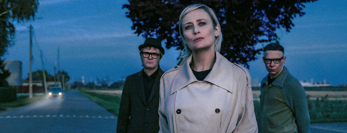 Drei Personen der Band Hooverphonic stehen auf einer Landstraße bei Dämmerung, vorne eine Person mit hellem Trenchcoat, dahinter zwei Personen mit dunkler Kleidung und Brillen