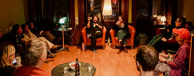 Mehrere Personen sitzen in einem Raum mit Holzfußboden, zwei Personen auf Sesseln in der Mitte, andere auf Stühlen im Halbkreis, Tisch mit Getränken im Vordergrund.