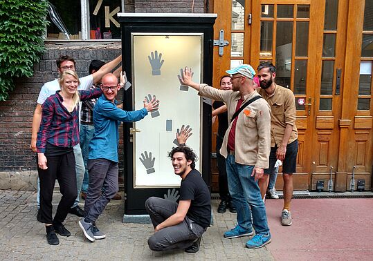 Gruppe von acht Personen vor einem hohen, schmalen Schaukasten mit mehreren schwarzen Handabdrücken auf weißem Hintergrund, einige Personen berühren die Handabdrücke.