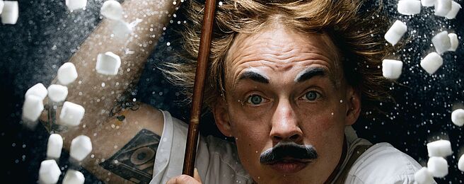 Person mit hochgekrempelten Ärmeln hält einen langen Stab, umgeben von fliegenden kleinen weißen Würfeln und weißem Pulver vor dunklem Hintergrund.