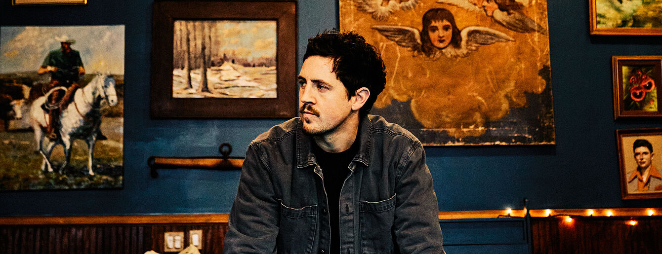 Michael Marcagi sitzt in einem Raum mit blauen Wänden, umgeben von verschiedenen Gemälden, und schaut nach links