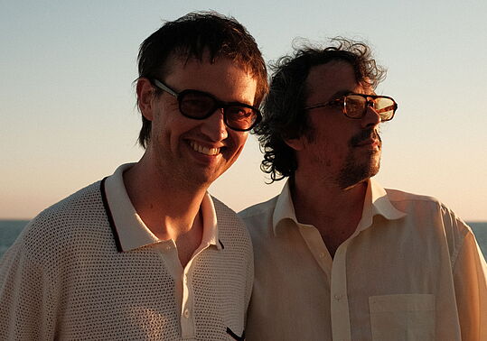 zwei männlich gelesene Personen stehen im Sonnenuntergang beim Meer mit Sonnenbrillen und grinsen in die Kamera