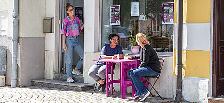 Foto: Susanne Senekowitsch zwei Personen sitzen an einem kleinen lila Tisch vor einem Café, eine weitere Person steht in der Tür. Bunte Girlande hängt im Fenster