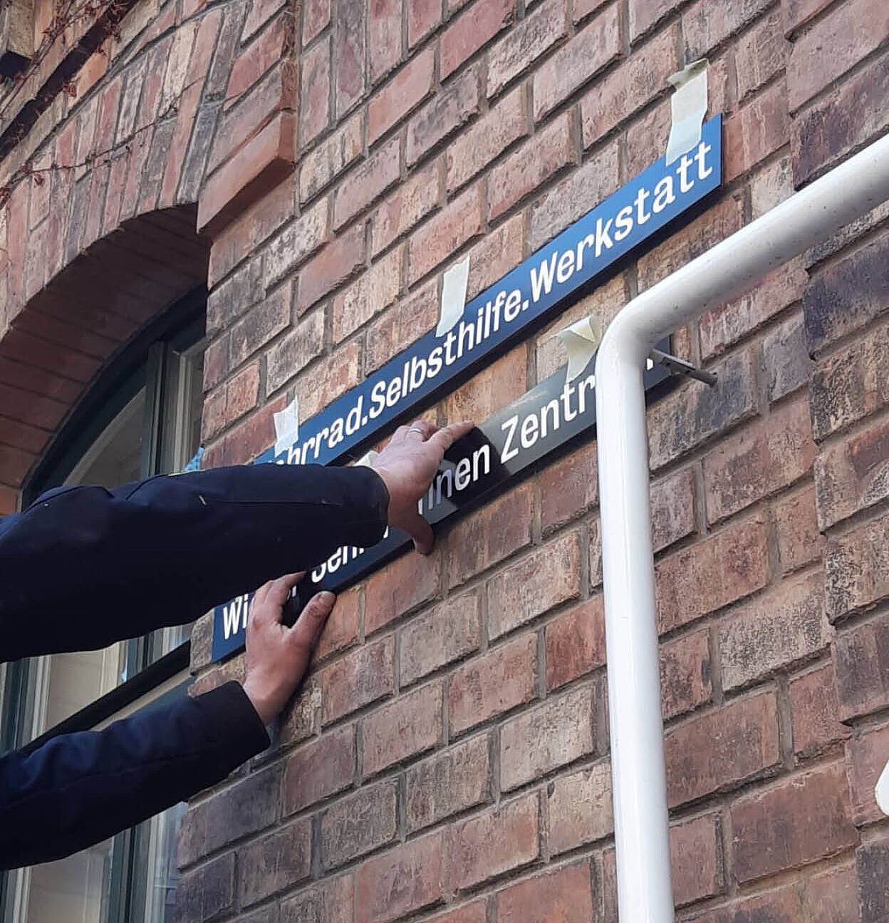 Eine Person mit dunkler Jacke befestigt ein blaues und ein schwarzes Wegweisschild mit weißer Schrift an einer Backsteinfassade. Die Schilder sind mit Klebestreifen provisorisch fixiert und zeigen den Namen verschiedener Einrichtungen.