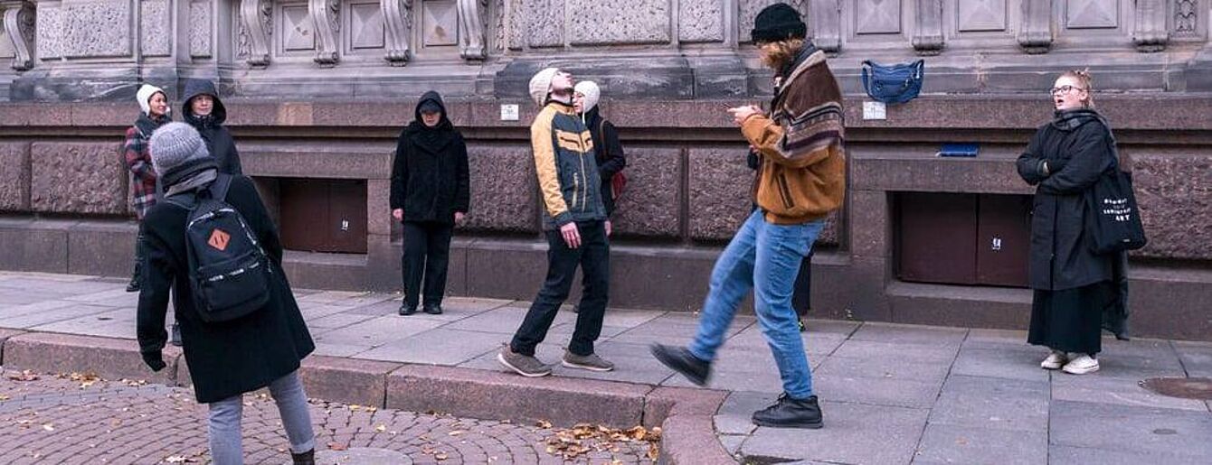 Außenaufnahme einer Gruppe von Menschen, die auf einem Gehweg vor einem großen, hellbeigen Steingebäude mit vielen Fenstern und komplizierten architektonischen Details stehen. Der Gehweg ist mit grauem und braunem Stein gepflastert. Ein kreisförmiger, rötlich-brauner Ziegelstein umrandet einen Fleck mit heruntergefallenen Blättern und Schutt. Auf dem Gehweg stehen oder gehen mehrere Menschen, die unterschiedlich gekleidet sind und verschiedene Tätigkeiten ausüben. Einige tragen Wintermützen und Jacken, andere tragen Mäntel und Jeans. Einige Personen scheinen sich Sehenswürdigkeiten anzusehen oder miteinander zu sprechen. Ein junger Erwachsener trägt eine hellbraune Jacke und Jeans und scheint ein Smartphone zu benutzen. Das Gebäude ist sehr hoch und breit, mit vielen Fenstern und mehreren Schichten von Mauerwerk. Das Gebäude weist eine Mischung aus hellbeigem und gräulichem Stein sowie regelmäßige, dekorative Muster auf. Der allgemeine Stil lässt auf ein formelles oder historisches Gebäude schließen. Die Beleuchtung erinnert an einen bedeckten Tag mit schwacher, aber klarer Sicht. Die Farben sind zurückhaltend, aber natürlich.