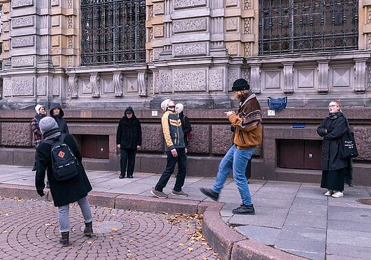 Außenaufnahme einer Gruppe von Menschen, die auf einem Gehweg vor einem großen, hellbeigen Steingebäude mit vielen Fenstern und komplizierten architektonischen Details stehen. Der Gehweg ist mit grauem und braunem Stein gepflastert. Ein kreisförmiger, rötlich-brauner Ziegelstein umrandet einen Fleck mit heruntergefallenen Blättern und Schutt. Auf dem Gehweg stehen oder gehen mehrere Menschen, die unterschiedlich gekleidet sind und verschiedene Tätigkeiten ausüben. Einige tragen Wintermützen und Jacken, andere tragen Mäntel und Jeans. Einige Personen scheinen sich Sehenswürdigkeiten anzusehen oder miteinander zu sprechen. Ein junger Erwachsener trägt eine hellbraune Jacke und Jeans und scheint ein Smartphone zu benutzen. Das Gebäude ist sehr hoch und breit, mit vielen Fenstern und mehreren Schichten von Mauerwerk. Das Gebäude weist eine Mischung aus hellbeigem und gräulichem Stein sowie regelmäßige, dekorative Muster auf. Der allgemeine Stil lässt auf ein formelles oder historisches Gebäude schließen. Die Beleuchtung erinnert an einen bedeckten Tag mit schwacher, aber klarer Sicht. Die Farben sind zurückhaltend, aber natürlich.