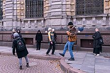 Außenaufnahme einer Gruppe von Menschen, die auf einem Gehweg vor einem großen, hellbeigen Steingebäude mit vielen Fenstern und komplizierten architektonischen Details stehen. Der Gehweg ist mit grauem und braunem Stein gepflastert. Ein kreisförmiger, rötlich-brauner Ziegelstein umrandet einen Fleck mit heruntergefallenen Blättern und Schutt. Auf dem Gehweg stehen oder gehen mehrere Menschen, die unterschiedlich gekleidet sind und verschiedene Tätigkeiten ausüben. Einige tragen Wintermützen und Jacken, andere tragen Mäntel und Jeans. Einige Personen scheinen sich Sehenswürdigkeiten anzusehen oder miteinander zu sprechen. Ein junger Erwachsener trägt eine hellbraune Jacke und Jeans und scheint ein Smartphone zu benutzen. Das Gebäude ist sehr hoch und breit, mit vielen Fenstern und mehreren Schichten von Mauerwerk. Das Gebäude weist eine Mischung aus hellbeigem und gräulichem Stein sowie regelmäßige, dekorative Muster auf. Der allgemeine Stil lässt auf ein formelles oder historisches Gebäude schließen. Die Beleuchtung erinnert an einen bedeckten Tag mit schwacher, aber klarer Sicht. Die Farben sind zurückhaltend, aber natürlich.