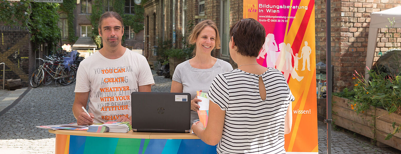 Zwei Beratungspersonen am Informationsstand der Bildungsberatung WUK in Wien sprechen mit Person Frau in einem Innenhof.