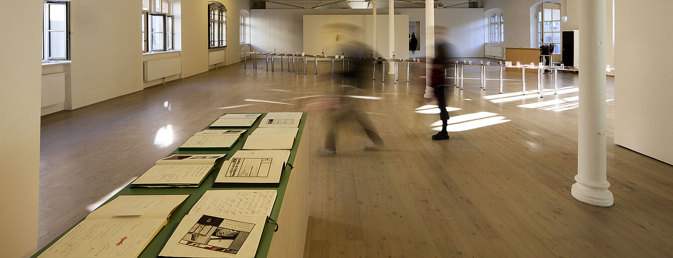 Großer Ausstellungsraum der kex mit Holzboden und hohen Fenstern. Auf einem langen Tisch liegen mehrere Gästebücher. Zwei Personen gehen durch den Raum. Die Personen sind unscharf.
