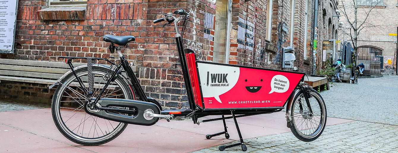 Schwarzes Lastenrad 'Grätzlrad' mit roter Ladefläche und Aufschrift 'WUK' und 'Du kannst mich ausborgen!' vor Backsteinmauer