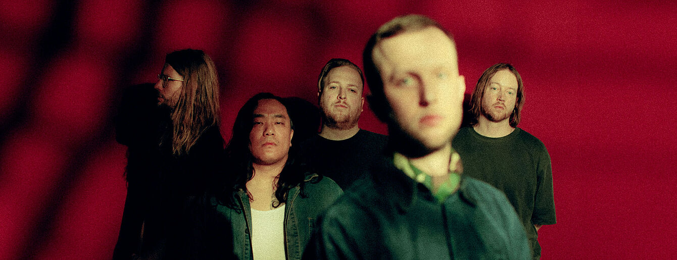 Fünf Mitglieder der Band La Dispute vor rotem Hintergrund, vorn unscharf ein blondes Bandmitglied, dahinter vier Mitglieder mit unterschiedlichen Frisuren und dunkler Kleidung