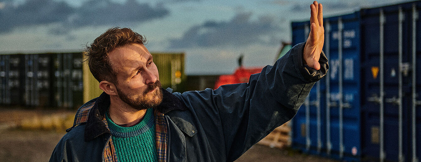 Florian Künstler steht zwischen Containern und blickt gegen die Sonne, mit seiner Hand versucht er das Sonnenlicht aus seinem Gesicht zu halten.