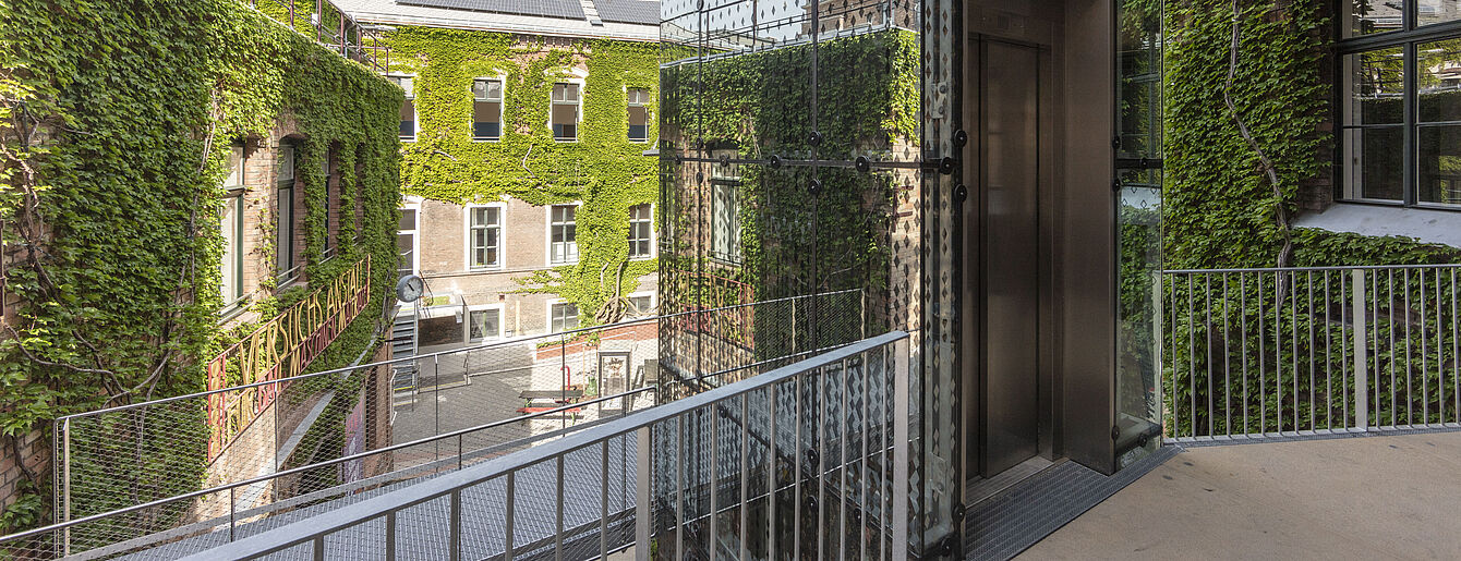 Blick auf einen Innenhof mit begrünten Wänden und einem gläsernen Aufzug. Links sind Pflanzen an den Wänden, rechts ein Gebäude mit Fenstern.