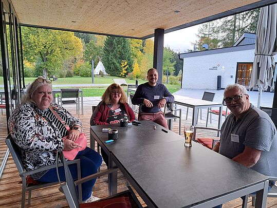 4 Personen sitzen draußen auf einem Tisch, im Hintergrund sieht man ein Haus und eine Wiese