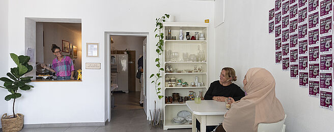 Zwei Frauen sitzen an einem weißen Tisch in einem hellen Raum mit weißen Wänden, an der Wand hängen viele kleine quadratische Bilder in einem Raster, im Hintergrund steht ein Regal mit Gläsern und Pflanzen, links ist eine Durchreiche mit einer Person dahinter.