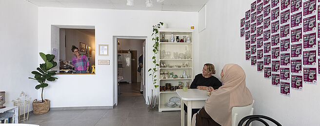 Zwei Frauen sitzen an einem weißen Tisch in einem hellen Raum mit weißen Wänden, an der Wand hängen viele kleine quadratische Bilder in einem Raster, im Hintergrund steht ein Regal mit Gläsern und Pflanzen, links ist eine Durchreiche mit einer Person dahinter.