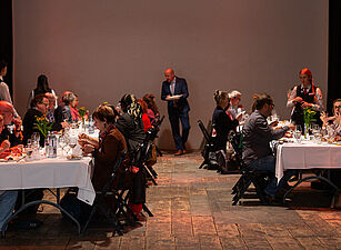 Foto: Susanne Senekowitsch Mehrere Personen sitzen an gegenüberstehenden, weiß gedeckten Tischen und essen, im Hintergrund steht ein Mann mit Notizblock.