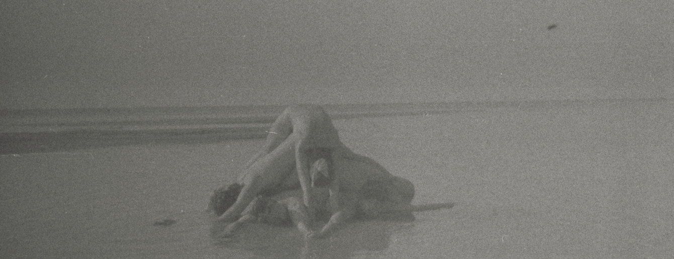 Full outdoor shot of a grayscale image, likely a vintage photograph or film frame. The foreground shows a beach or sandy area. Several indistinct human figures are clustered together, lying or positioned in a way that suggests activity of some kind. The figures lack clear detail, appearing as light or dark shapes against the surrounding sand or water. The scene is somewhat blurred or grainy with a noticeable texture, typical of older photography or scans of film. The overall tonal range is muted, consisting primarily of grays, with subtle variations in shading. No distinct coloration is present. The background is a similar muted gray tone extending out of view, lacking any contrasting light or shadow that would make the figures especially noticeable.