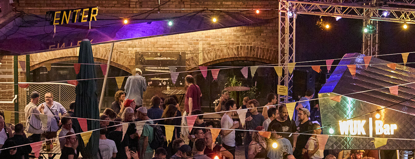 Menschenmenge bei einer Veranstaltung im WUK mit beleuchtetem Schriftzug 'WUK | Bar' und bunten Wimpelketten