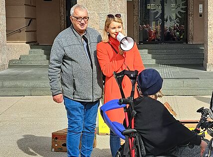 2 Personen stehen vor einem Gebäude, eine hält ein Megaphon, davor sitzt eine Person im Rollstuhl