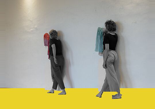 Zwei Frauen mit lockigem Haar stehen barfuß vor einer weißen Wand, jede hält ein Handtuch, eine rote und eine blaue, auf gelbem Boden