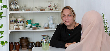Foto: Susanne Senekowitsch Zwei Frauen sitzen an einem weißen Tisch, im Hintergrund ein Regal mit Keramikgeschirr und Gläsern