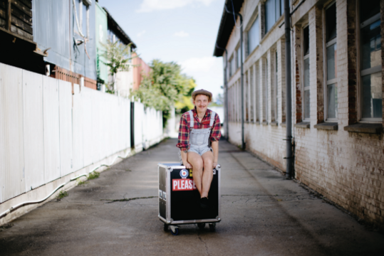 Person in Latzshorts und kariertem Hemd sitzt auf einem schwarzen Rollkoffer in einem engen Hof zwischen weißen und ziegelsteinernen Gebäuden