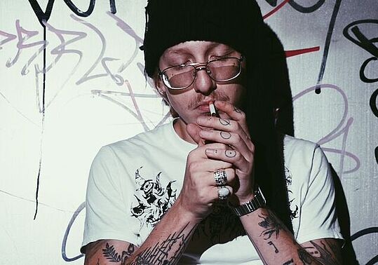 Edo Saiya am 15.4.2026 im WUK Wien - Pressefoto von Edo Saiya, der mit Mütze aus, an Wand angelehnt sitzend eine Zigarette anzündet