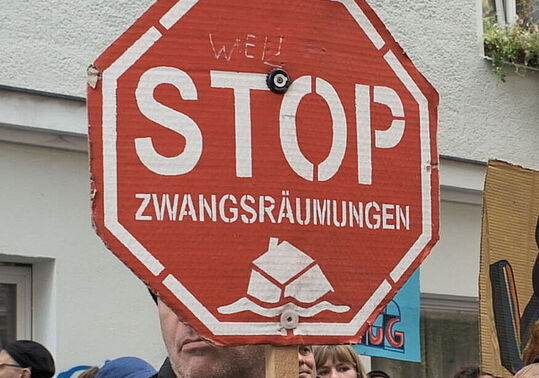 Weiblich und männlich gelesene Demonstrant*innen halten rote Stoppschilder mit der Aufschrift 'STOP ZWANGSRÄUMUNGEN' vor einem weißen Gebäude mit Fenstern.