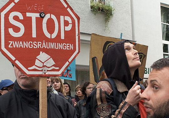 Weiblich und männlich gelesene Demonstrant*innen halten rote Stoppschilder mit der Aufschrift 'STOP ZWANGSRÄUMUNGEN' vor einem weißen Gebäude mit Fenstern.